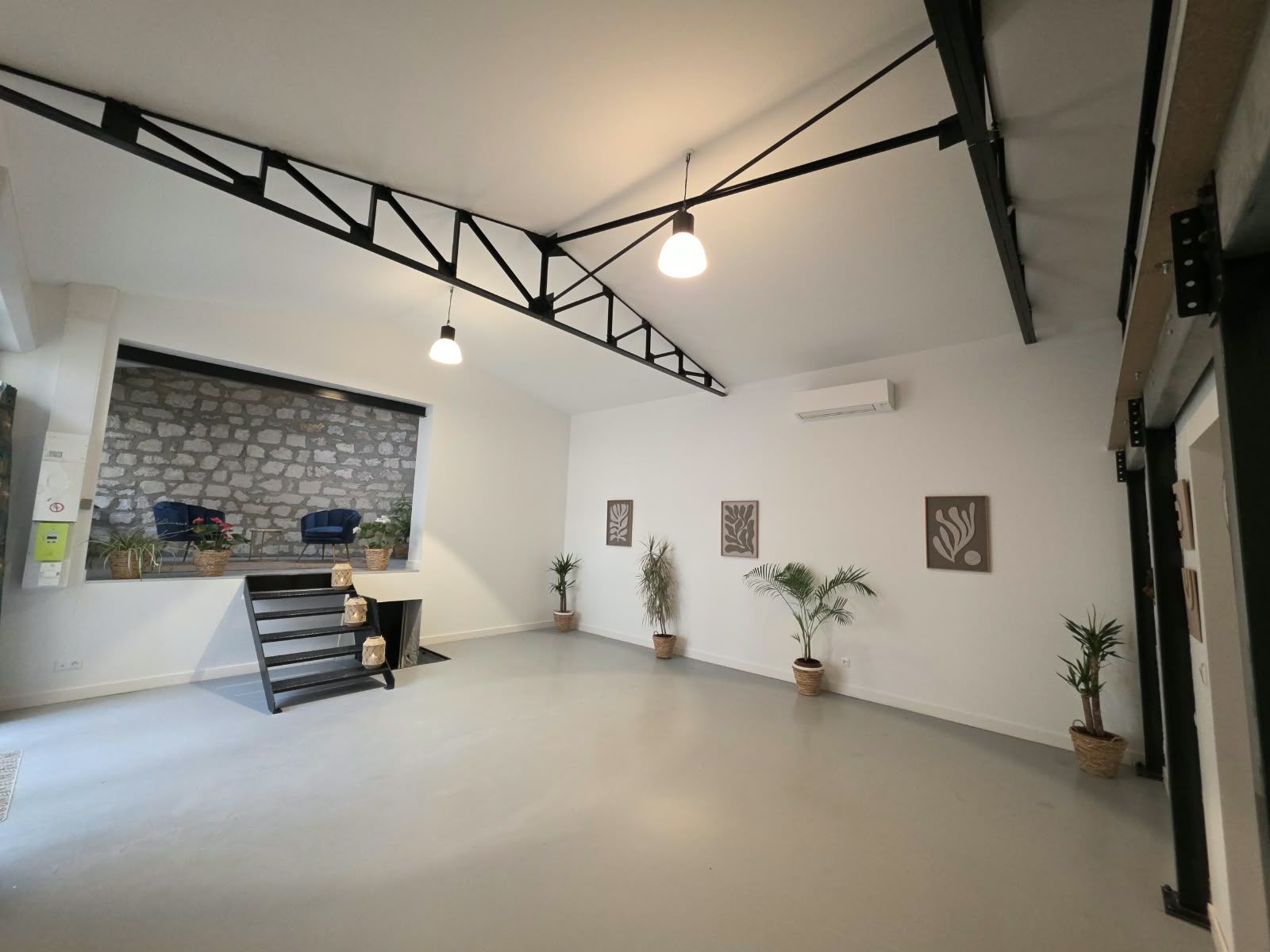 La salle vide de l’Institut Sãdhanã : grand espace clair sous une charpente métallique noire, plantes, et l’estrade au mur de pierre au fond